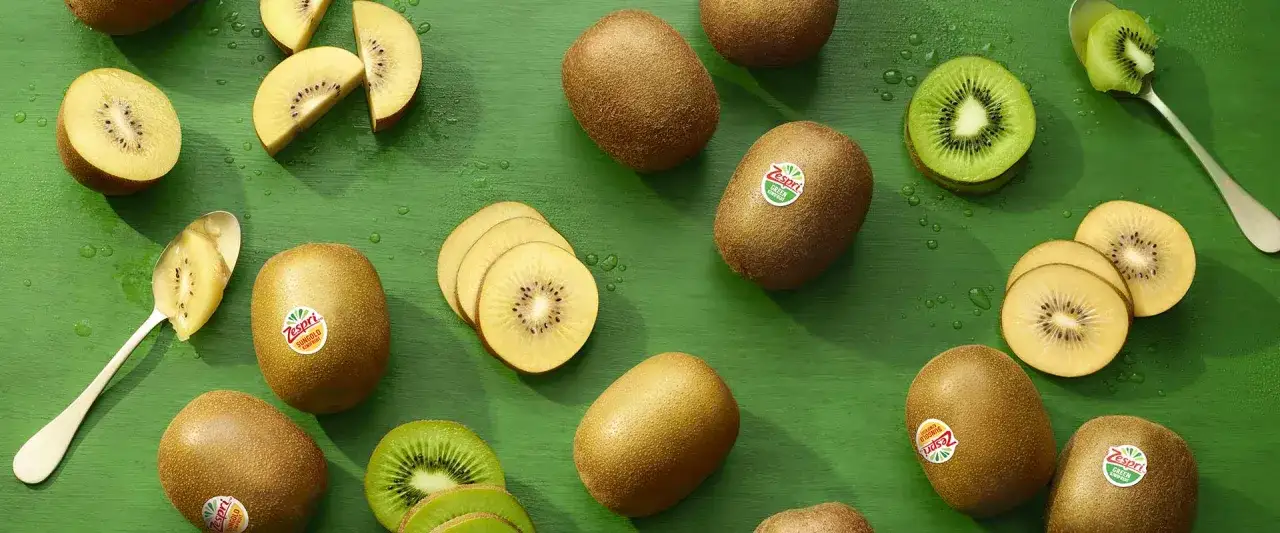 Goldene und gr&uuml;ne Kiwis, einige ganz, andere aufgeschnitten, liegen auf einem gr&uuml;nen Untergrund. Ein L&ouml;ffel mit Kiwi-Fruchtfleisch. Ein s&uuml;&szlig;er Traum vor dem Schlafen.