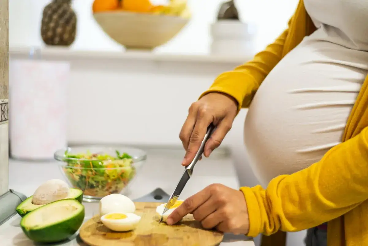Schwangere bereitet gesunde Mahlzeit zu. Eier in der Schwangerschaft sind wichtig f&uuml;r die N&auml;hrstoffversorgung. Avocado und Salat sind ebenfalls zu sehen.